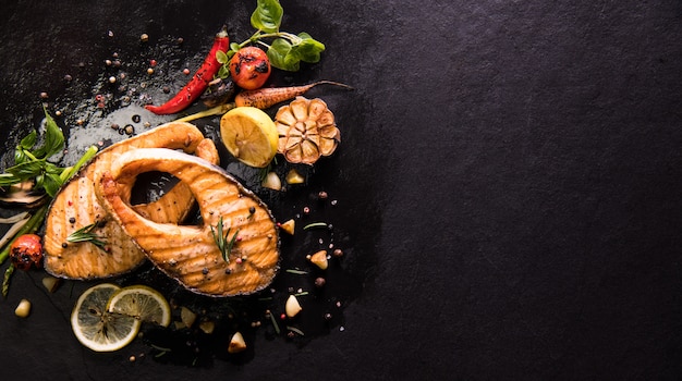 Grilled salmon fish with seasoning and various vegetables on black stone background