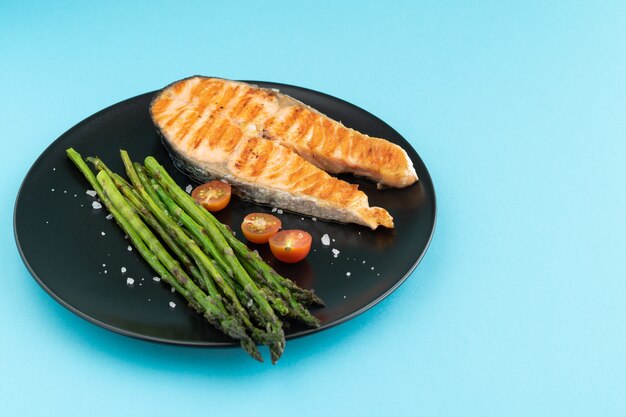 Grilled salmon slice with green asparagus on black plate on blue background. copy space.