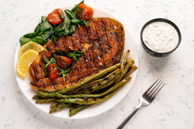 Grilled salmon steak, asparagus and fresh spinach with tomatoes on a white plate on a light table.