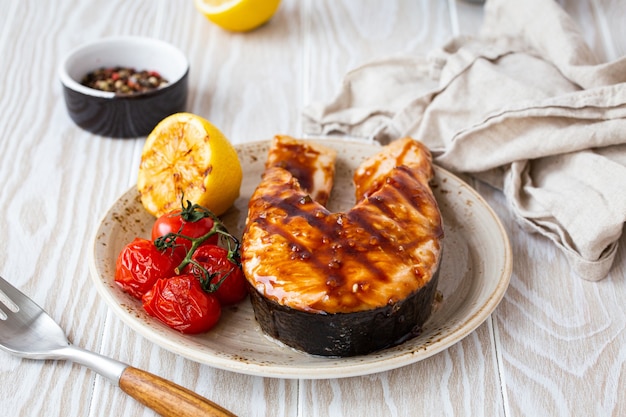 Grilled salmon steak glazed with teriyaki sauce, vegetables and lemon served on ceramic plate on rustic white wooden table from angle view, selective focus