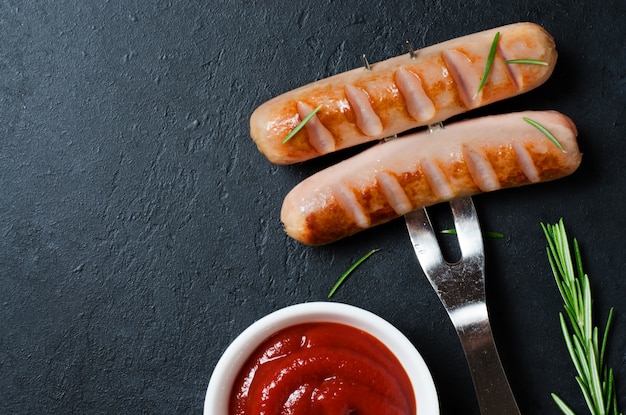 Grilled sausages fried on a metal fork. Ketchup, rosemary. Unhealthy diet. Dark background