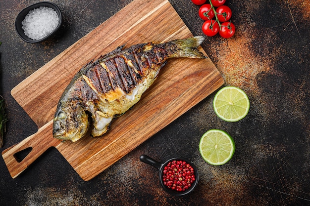 Grilled sea bream or dorado raw fish on chopping  board with ingredients over old metall dark background, top view with space for text.