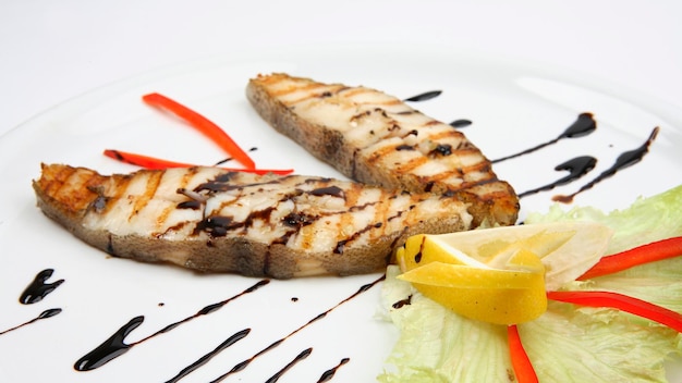 Grilled seafood halibut with lemon, red pepper and green lettuce on white plate. Close-up photo