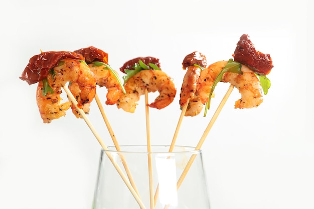 Grilled shrimps with the dried tomato and arugula on sticks, a canape stand in a glass on a white background