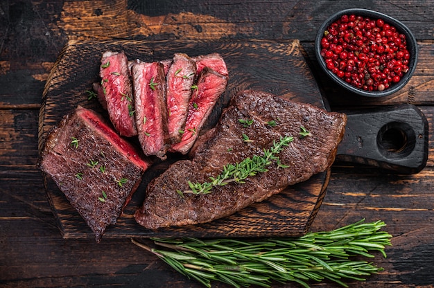 Grilled sliced skirt beef meat steak on a cutting board with herbs. Dark wooden background. Top view.