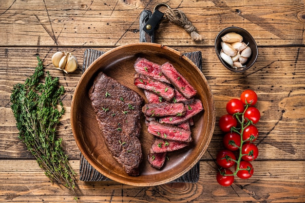 Grilled sliced top blade or denver beef meat steak in a wooden plate with herbs. wooden background. Top view.