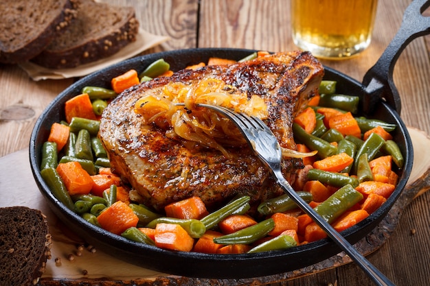 Bistecca alla griglia da carne di maiale con cipolle fritte, fagiolini e carote in una padella su un fondo di legno naturale.