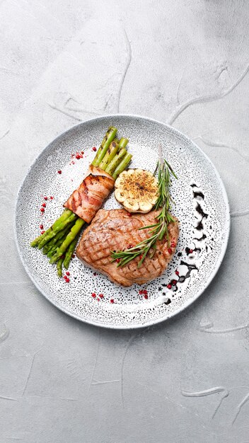 Photo grilled steak with asparagus and rosemary leaf in plate top view on concrete background with copy space.
