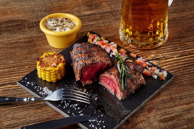 Grilled steak with corn with mushrooms sauce on cutting board and mug of beer on wooden table