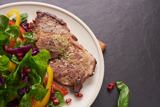 Bistecca alla griglia con verdura fresca, peperone dolce, pomodori, cipolla rossa, pepe rosa e spezie. cibo gustoso fatto in casa. concetto per un pasto gustoso e sano. superficie in pietra nera. bistecca di maiale con insalata