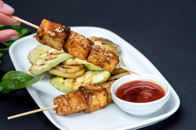 白い皿に豆腐ケバブを焼きました。黒の背景。新鮮なバジルの葉。