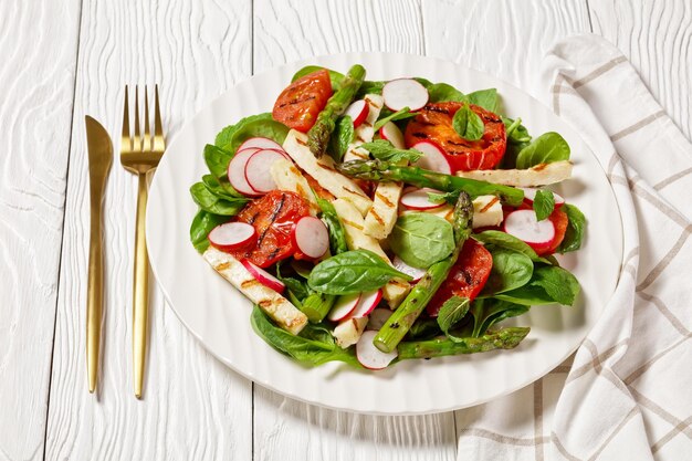 Pomodori grigliati, formaggio halloumi, asparagi, ravanelli e insalata di spinaci su un piatto su un tavolo di legno bianco, vista orizzontale dall'alto