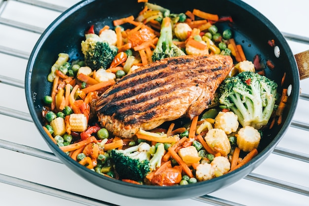 Photo grilled turkey steak fillet and fried vegetables mix in a black pan on the white table.