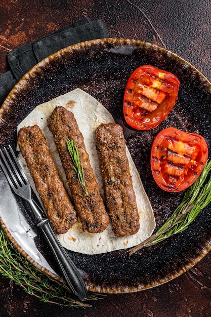 Grilled urfa shish kebab on a plate with tomato dark background\
top view