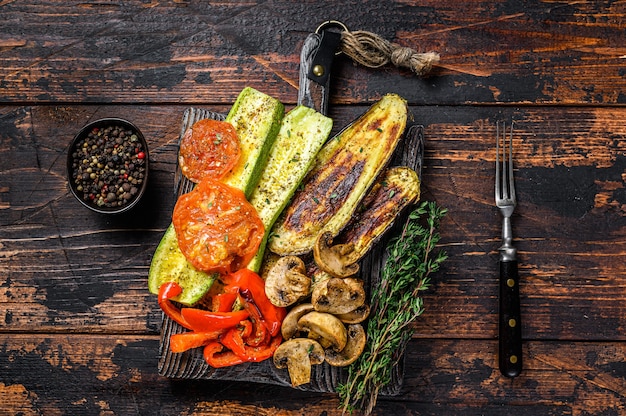 Grilled vegetables bell pepper, zucchini, eggplant and tomato with dry herbs on a wooden board. Dark wooden background. Top view.