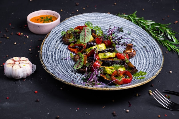 Grilled vegetables zucchini, eggplant, mushrooms, bell peppers combined with green butter in a blue plate, on a dark surface