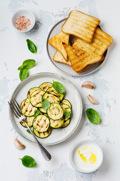 Grilled zucchini salad with basil leaves, yogurt sauce and fried bread in a simple ceramic plate on a white concrete surface