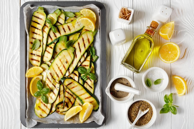 Grilled zucchini slices on a sheet pan with lemon, olive oil, spices and fresh basil leaves on a white wooden table, flat lay, italian cuisine