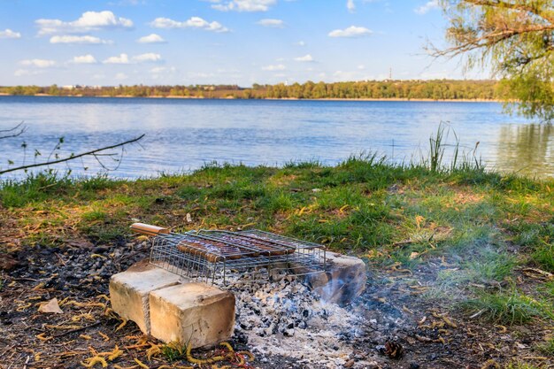 Grilling sausages in portable barbecue grill on campfire at riverbank