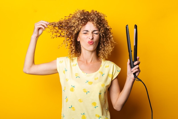 La giovane donna smorfia raddrizza i capelli ricci con una piastra per capelli su uno sfondo giallo.