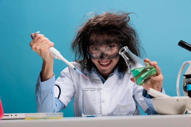 Grinning creepy lunatic silly looking mad scientist with dropper and beaker mixing experimental liquid chemical substances. Crazy chemist with dreadful face expression doing laboratory work.