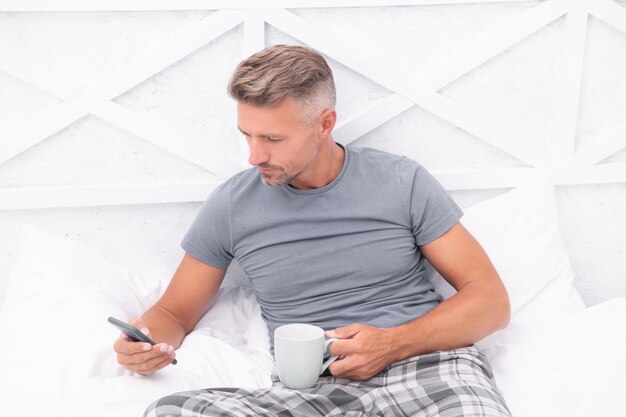Grizzled man online chatten in mobiele telefoon terwijl het drinken van koffie in bed Mobiele communicatie