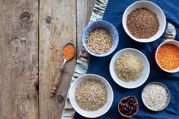 Groats in bowls on a wooden table.Linen tablecloth with dry cereals.Raw buckwheat, lentils, rice, beans, oatmeal, dry peas.View from above. Healthy food.Proper nutrition.Food concept.Vegan food.