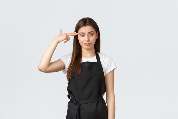 Grocery store employees, small business and coffee shops concept. Annoyed and tired female barista in black apron pointing fake gun at head irritated, staring skeptical and bothered