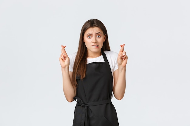 Grocery store employees, small business and coffee shops concept. Anxious cute female barista in black apron biting lip worried and cross fingers, praying or making wish.