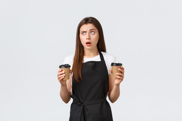 Grocery store employees, small business and coffee shops concept. Confused female barista with two cups of drinks looking upper left corner puzzled with opened mouth and frowning face