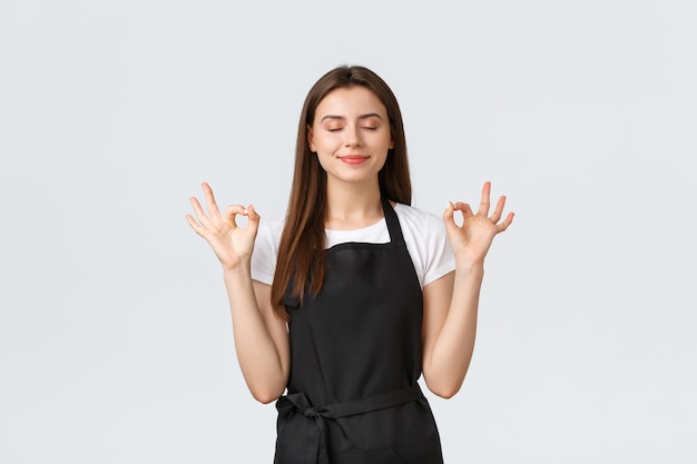 Grocery store employees, small business and coffee shops concept. Delighted cute smiling barista close eyes and smiling happy. Carefree cafe worker meditating, showing okay gesture.