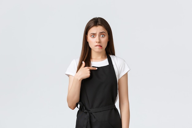 Grocery store employees, small business and coffee shops concept. Embarrassed and worried female barista in black apron biting lip and looking anxious, pointing herself. Copy space