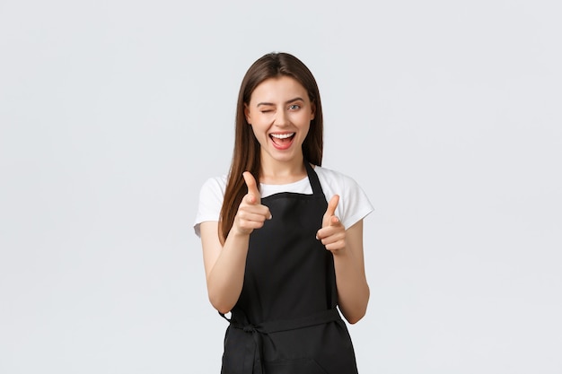 Dipendenti del negozio di alimentari, piccole imprese e concetto di caffetterie. emozionato barista sfacciato in grembiule nero che punta la fotocamera con le dita, si congratula con la persona, dicendo che ha suonato bene.