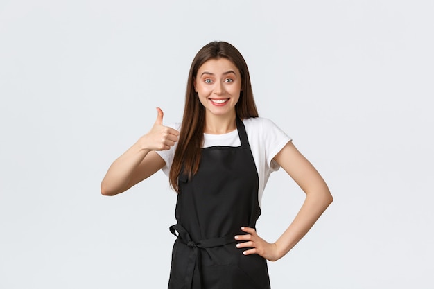 Grocery store employees, small business and coffee shops concept. Excited cheerful female barista show thumbs-up in approval. Saleswoman in black apron recommending product or special drink