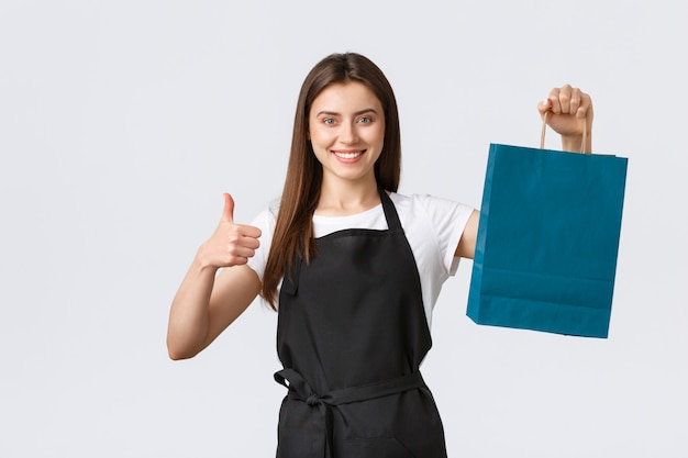 Grocery store employees, small business and coffee shops concept. Friendly cute saleswoman packing your gift or purchased items in paper bag, showing thumbs-up and smiling