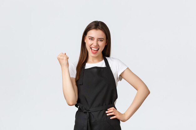 Grocery store employees, small business and coffee shops concept. Happy female barista in black apron fist pump in rejoice, celebrating great achievement or success