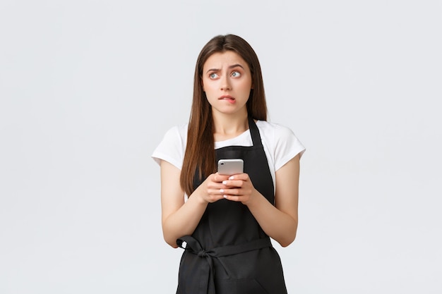 Grocery store employees, small business and coffee shops concept. Indecisive puzzled cute girl barista in black apron, looking left unsure, biting lip and using mobile phone, not sure what answer.