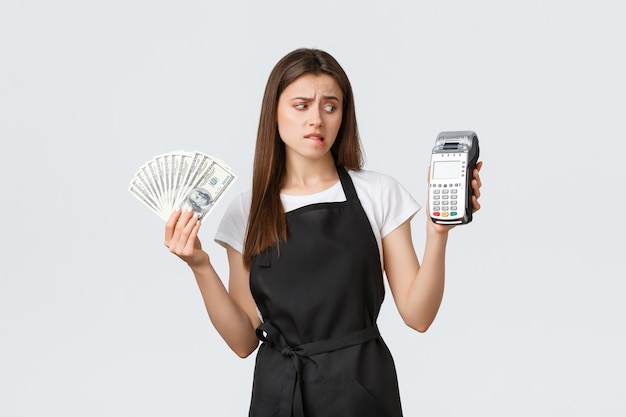 Grocery store employees, small business and coffee shops concept. Indecisive and unsure cute female barista showing cash and POS terminal, dont know how use it, stare concerned