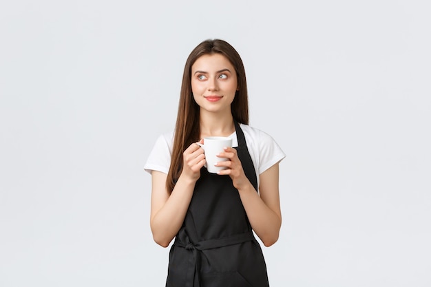 Grocery store employees, small business and coffee shops concept. Pleased cute saleswoman in black apron enjoying break, drinking tea and looking away with happy smile.