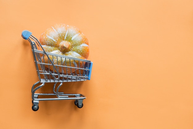 Grocery trolley with pumpkin on orange. Halloween shopping and sale. Flat lay, top view.