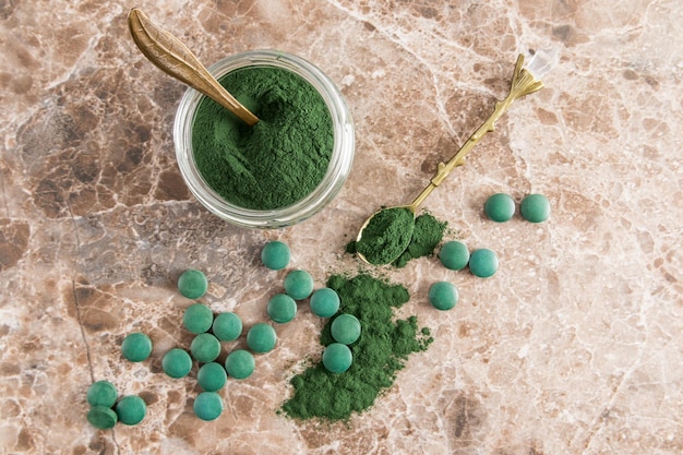 Groen spirulina-poeder in een glazen pot op een marmeren bruine achtergrond met verspreide spirulina-tabletten bovenaanzicht een gezond organisch supplement