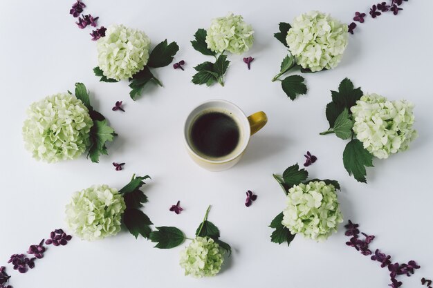 Groene bloemen en bloemblaadjes van lila in de vorm van een cirkel liggen rond de gele Cup met koffie op witte achtergrond