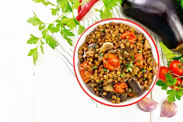 Groene linzen gestoofd met aubergine, tomaten, knoflook en kruiden in een kom op een theedoek, peterselie op houten plank achtergrond van bovenaf