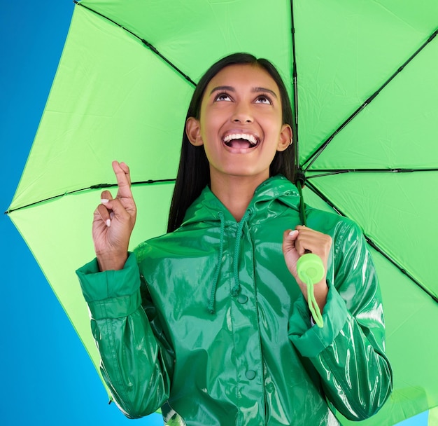 Foto groene paraplu en vingers gekruist met een vrouw op een blauwe achtergrond in de studio tijdens de winter voor verzekering regen mode of dekking en een aantrekkelijke jonge vrouw wensen voor het weer te veranderen met hoop