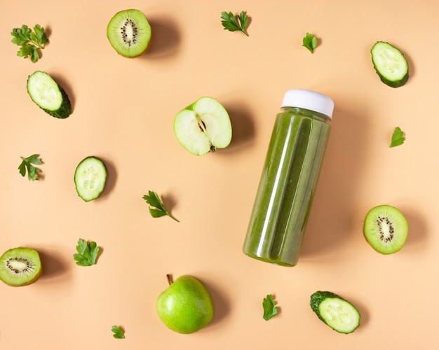 Groene smoothie in een transparante fles op een beige achtergrond. De gesneden groenten en fruit liggen klaar. Gezond eten, plat gelegd.