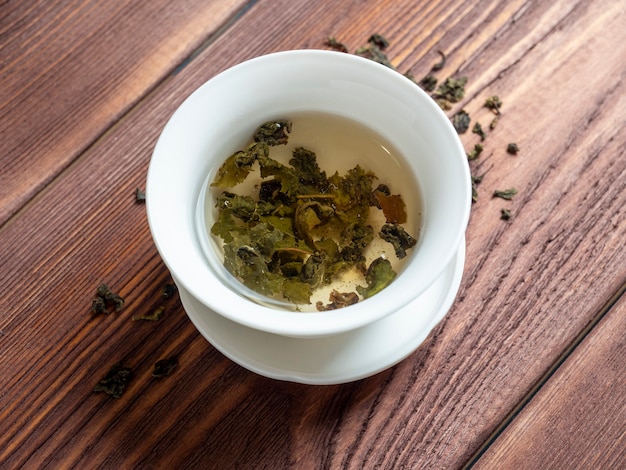 Groene thee wordt gebrouwen in een witte kom op een houten ondergrond. Droge theeblaadjes liggen vlakbij. Bovenaanzicht