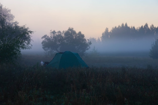 Groene toeristentent bij de rivier bij zonsopgang, met ochtendherfstmist op het water. Buiten toeristisch landschap.