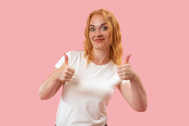 Groene vrouw met duimen omhoog gebaar op roze achtergrond kijkt in de camera Portret van jonge glimlachende ronde vrouw met roodharig haar
