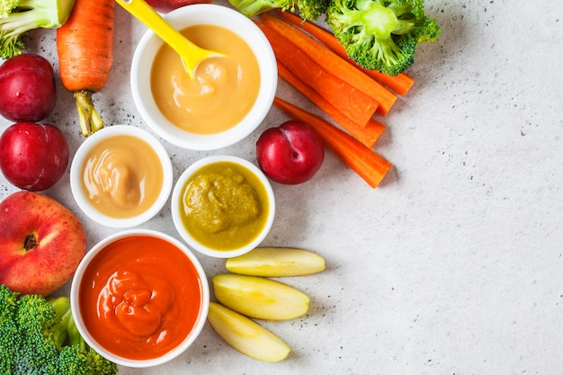 Groente- en fruitbabypuree in witte kommen met ingrediënten, bovenaanzicht.