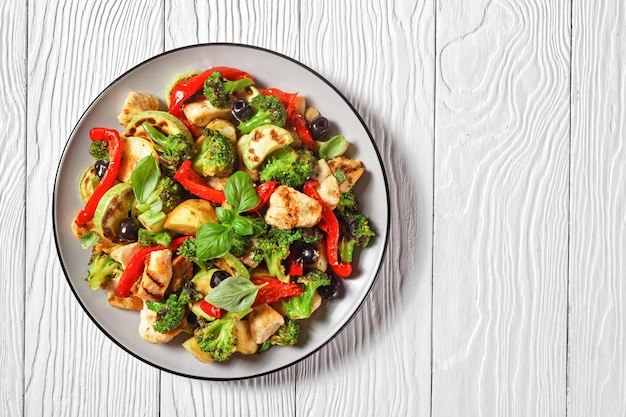 Groente- en kipgebraad met jonge aardappel, paprika, courgette, olijven, besprenkeld met balsamicoazijn en basilicum erop, geserveerd op een bord op witte houten ondergrond, bovenaanzicht, kopieerruimte